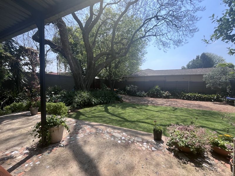 A covered patio area featuring mosaic tile flooring, overlooking a well-maintained garden with mature trees and perimeter walling.
