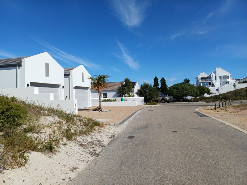 A clear street view of a residential neighbourhood featuring modern white homes with garages and paved access roads under a bright blue sky.