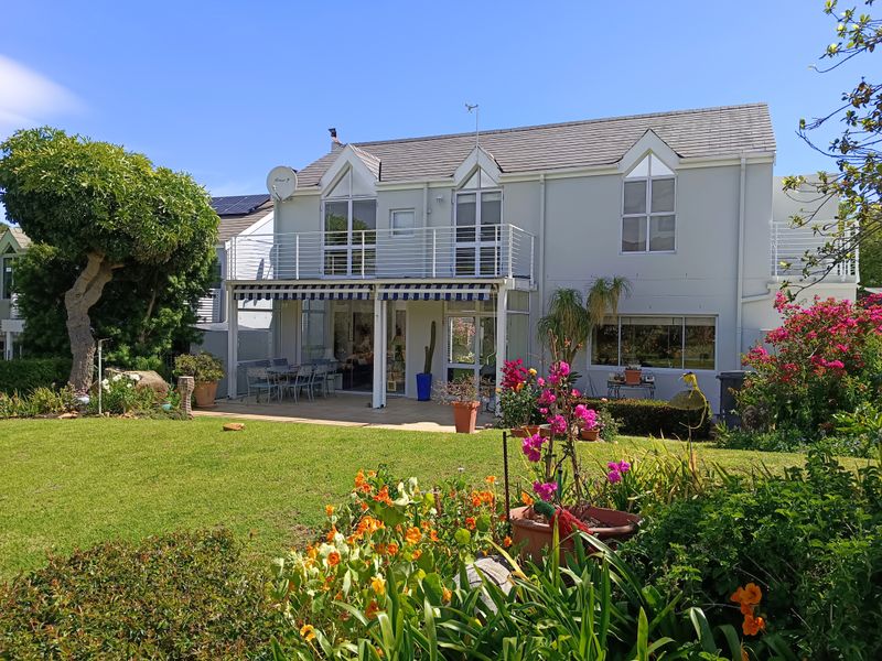Rear view of a two-story house featuring a paved patio, a balcony with metal railings, a retractable awning, and a well-maintained garden.