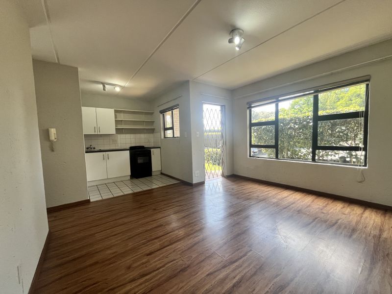 Open-plan living area featuring laminate flooring, large windows for natural light, and a compact kitchen with built-in cabinetry and a security gate.