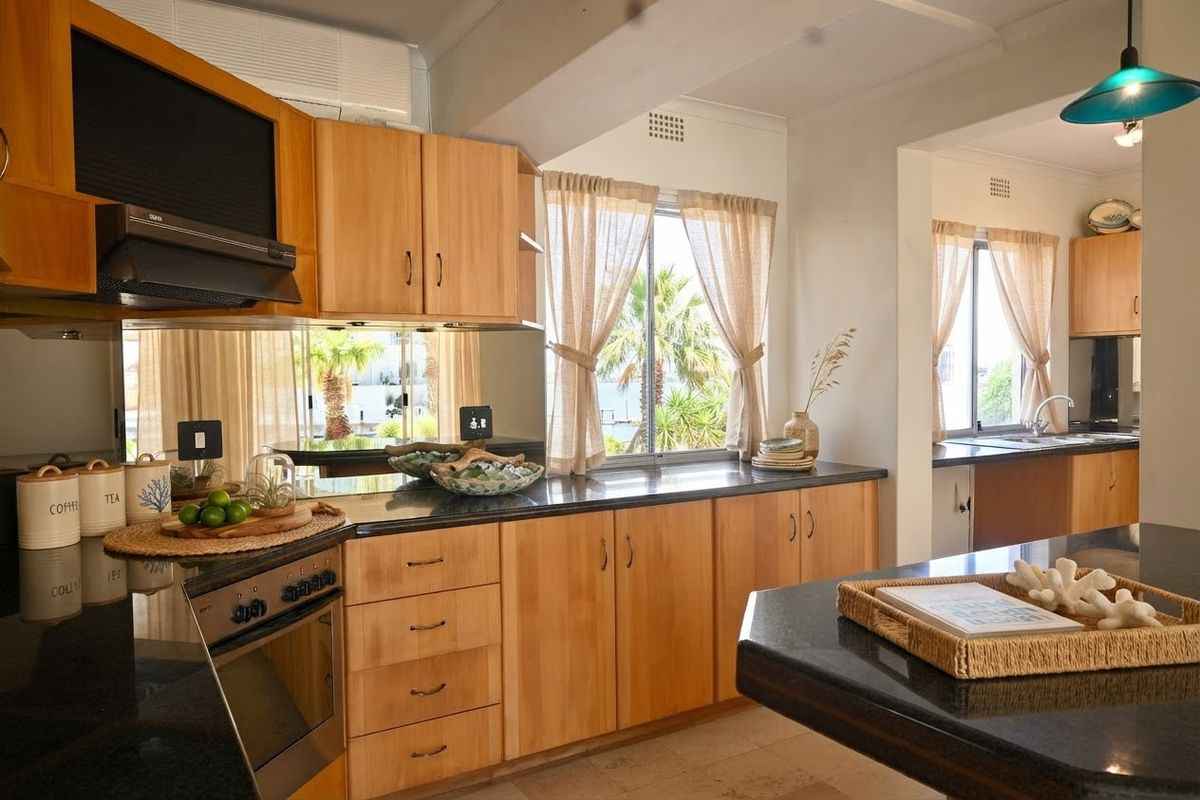 A well-maintained kitchen featuring warm wooden cabinetry, dark granite countertops, a mirrored backsplash, and a functional layout with an adjoining scullery area.