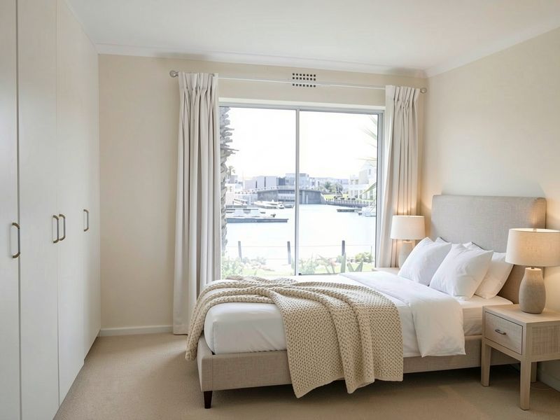 A bright bedroom featuring built-in wardrobes, neutral carpet flooring, and a sliding glass door offering scenic water views.