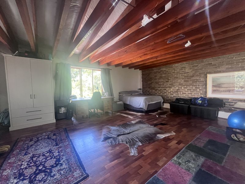 Spacious bedroom featuring exposed timber ceiling beams, a rustic brick feature wall, and warm hardwood flooring with natural light from a large window.