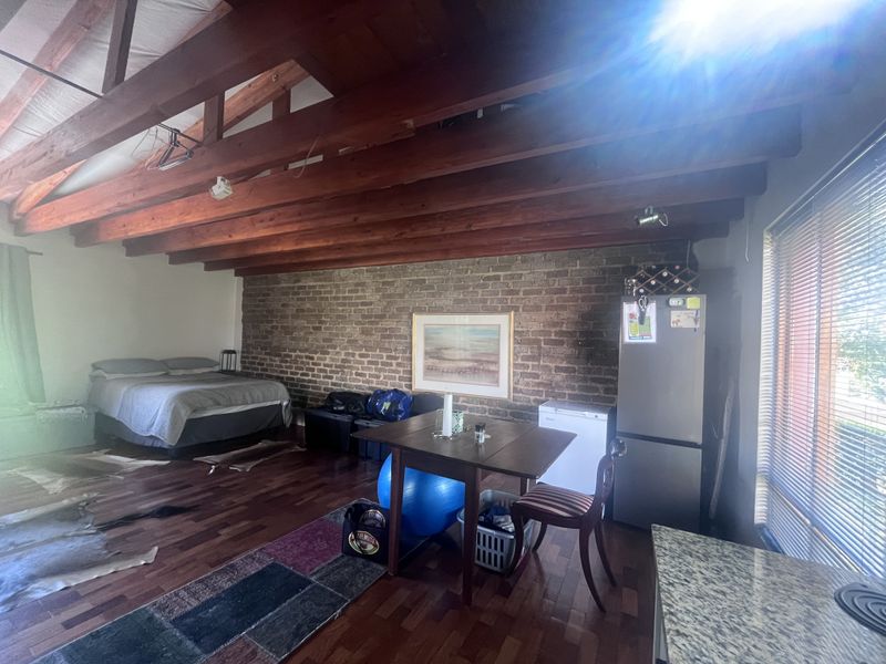 Open plan living area featuring exposed timber ceiling beams, a rustic brick feature wall, and hardwood flooring with large windows and venetian blinds.