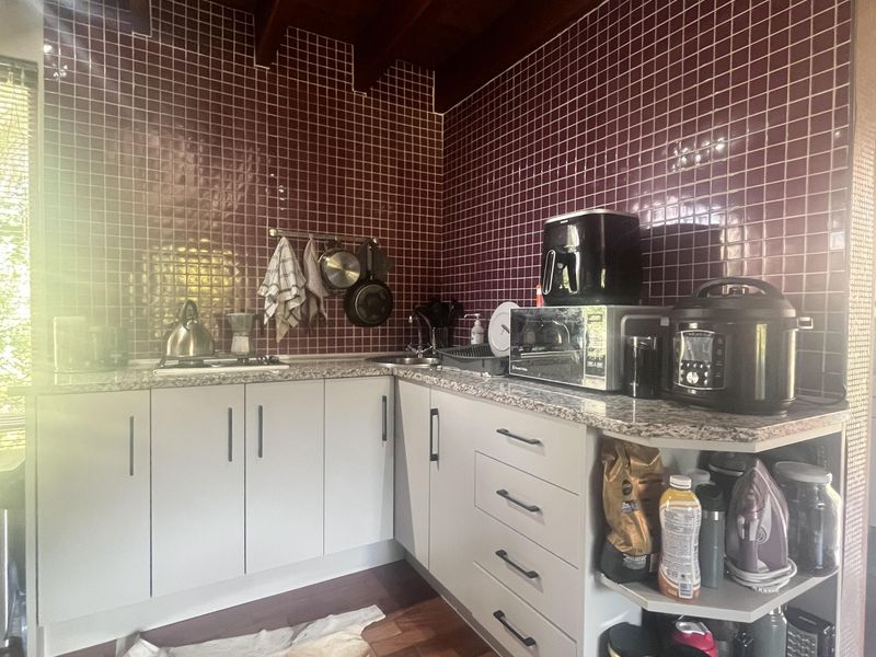 A compact kitchen featuring white cabinetry, granite-look countertops, a distinctive dark red tiled backsplash, and exposed timber ceiling beams.
