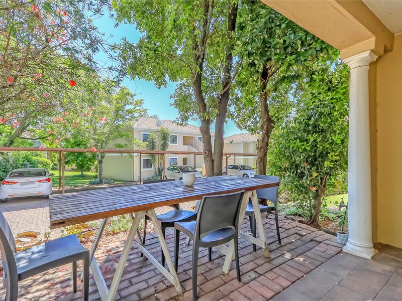 A covered patio area featuring paved flooring and a structural column, overlooking a landscaped garden and parking area.