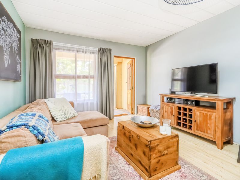 A comfortable living room featuring light laminate flooring, soft blue-toned walls, and a window providing natural light, leading to an adjacent room.