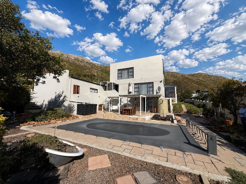Rear exterior view of the house featuring a paved patio area, swimming pool with cover, and a water tank set against a mountain backdrop.