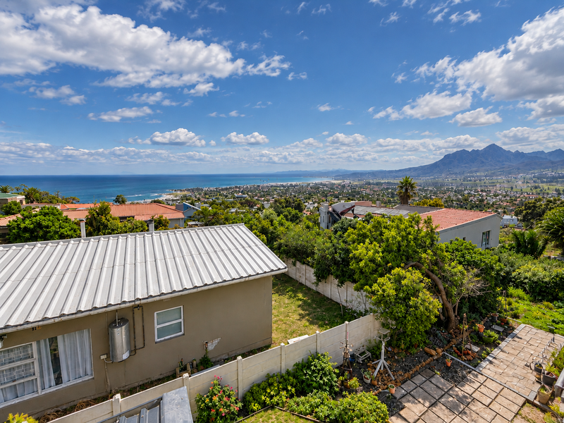Elevated view overlooking the residential neighborhood, coastline, and distant mountains, featuring a property with a corrugated metal roof and paved garden area.