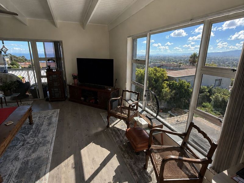 Bright living room featuring exposed beam ceilings, laminate flooring, and expansive windows offering panoramic views with access to an outdoor balcony.