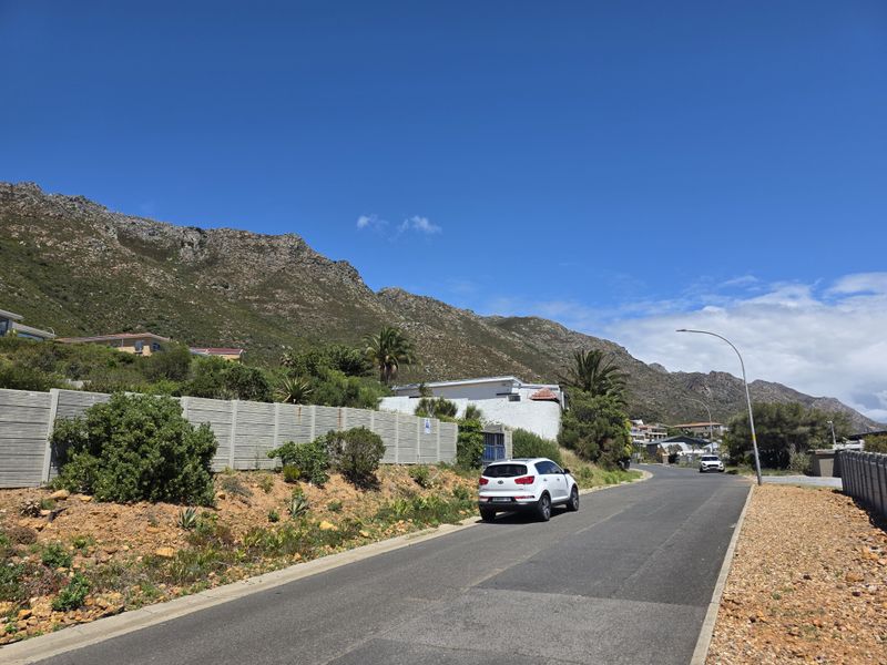 A street view showing the neighbourhood approach with a paved road, concrete boundary wall, and scenic mountain backdrop.