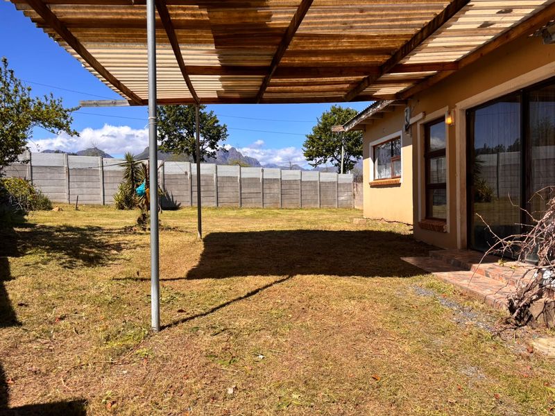 A covered outdoor patio area featuring a corrugated roof structure, sliding glass door access, and a spacious backyard enclosed by a precast concrete wall.