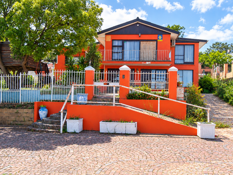 Front elevation of a two-story house featuring a vibrant orange facade, balcony, paved driveway, and accessible ramp entry.