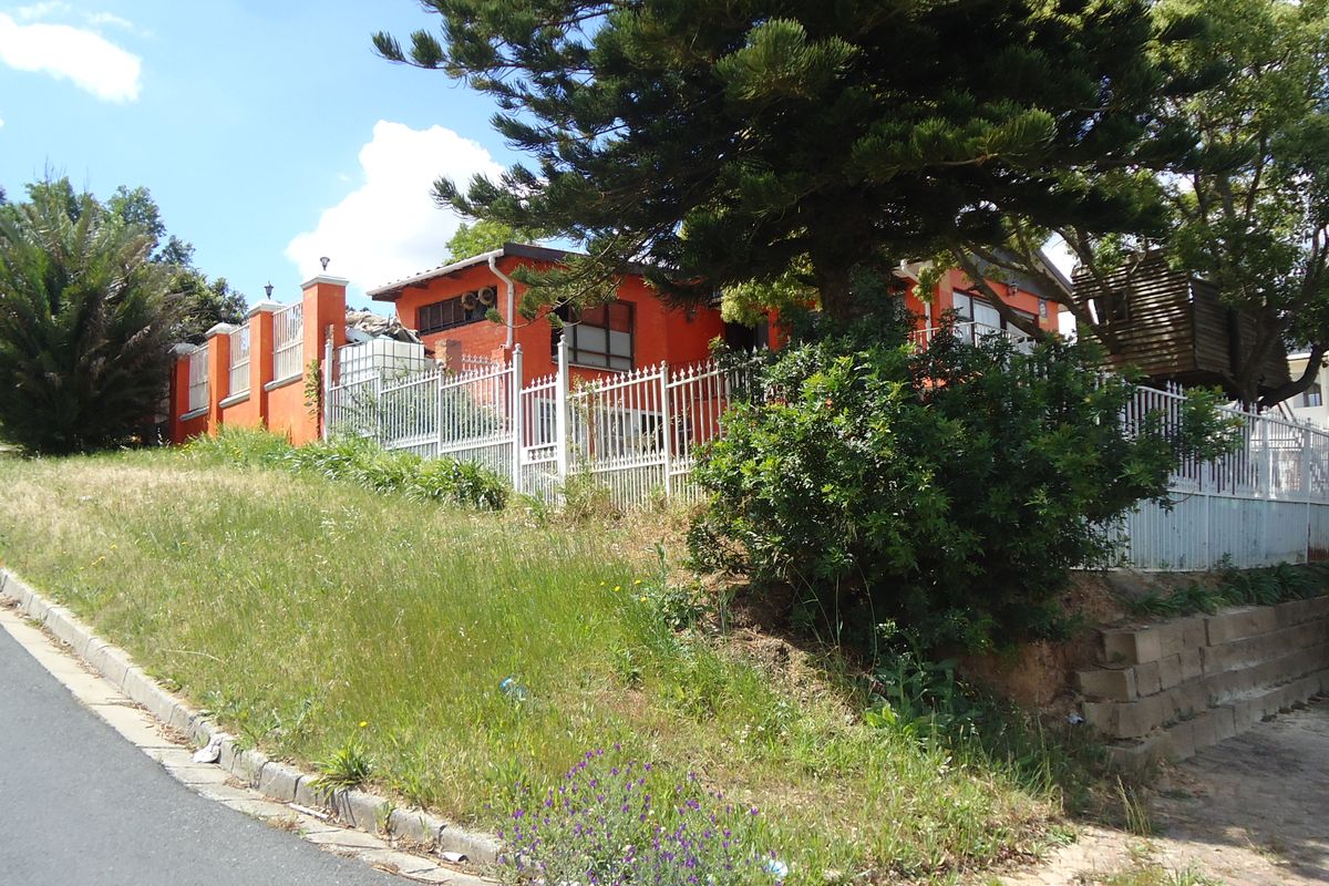 Street-level view of the property exterior featuring a bright orange facade, metal perimeter fencing, and a sloped garden area with mature trees.