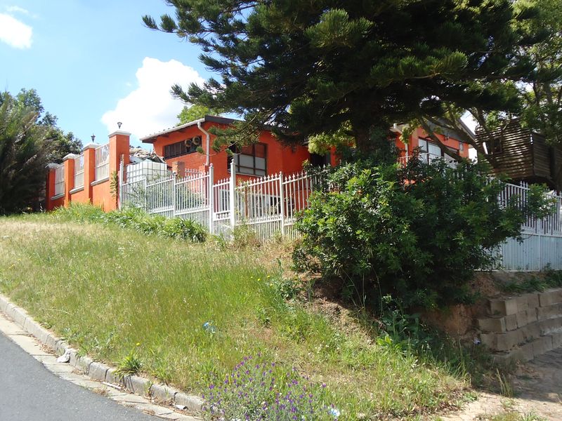 Street-level view of the property exterior featuring a bright orange facade, metal perimeter fencing, and a sloped garden area with mature trees.