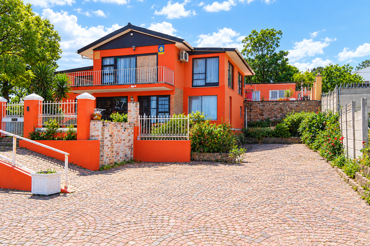 Front exterior view of a two-story house featuring a cobblestone driveway, metal fencing, and a balcony, set within a landscaped residential property.