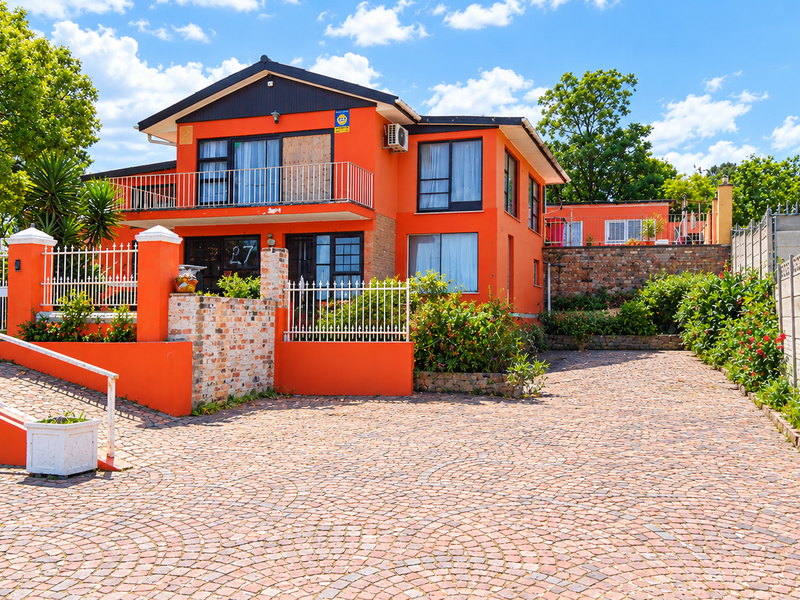 Front exterior view of a two-story house featuring a cobblestone driveway, metal fencing, and a balcony, set within a landscaped residential property.