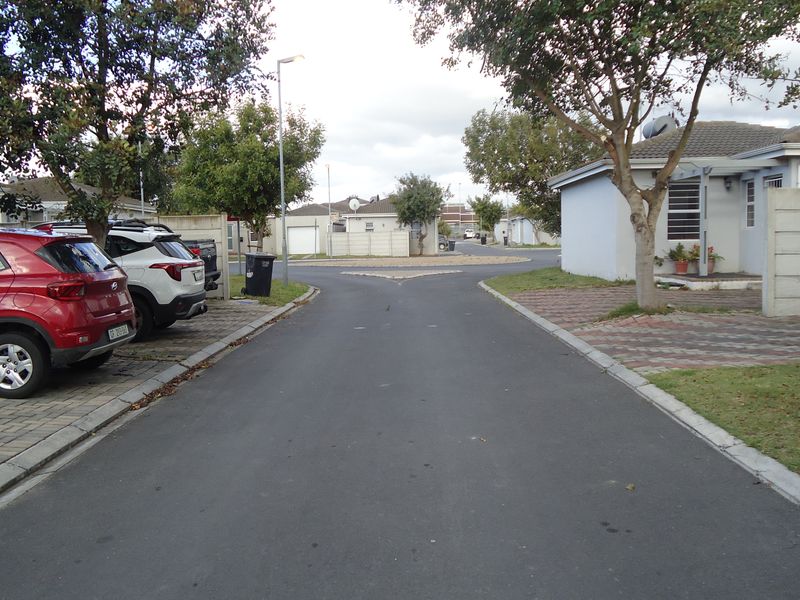 A view of a residential street within a housing complex, featuring paved driveways, boundary walls, and established trees.