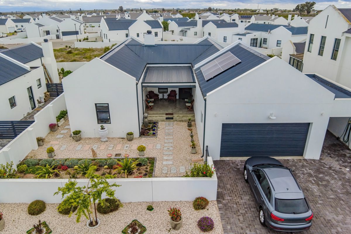 Aerial view of a modern house featuring a metal roof with solar panels, a paved driveway, a covered patio, and a well-maintained landscaped garden.