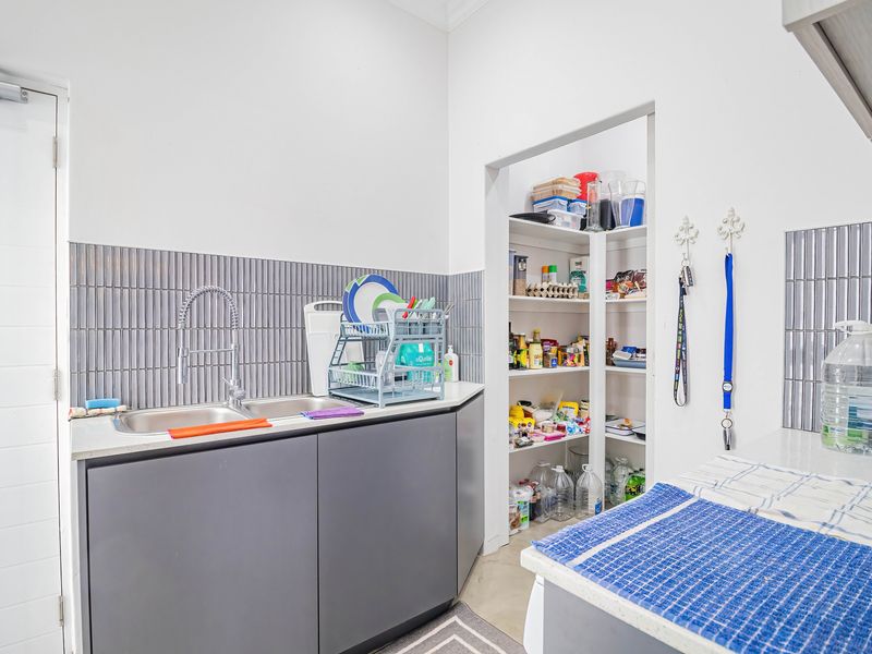A functional scullery featuring a double stainless steel sink, modern grey cabinetry, tiled splashback, and a convenient walk-in pantry with built-in shelving.