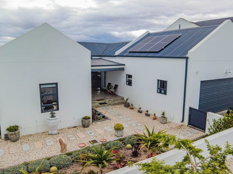 An elevated view of the front facade featuring a modern white exterior, metal roofing with integrated solar panels, and a landscaped gravel garden.
