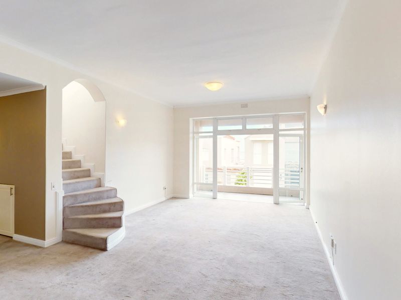 A bright, empty living room featuring neutral carpeting, an arched staircase, and large glass doors leading to a balcony.