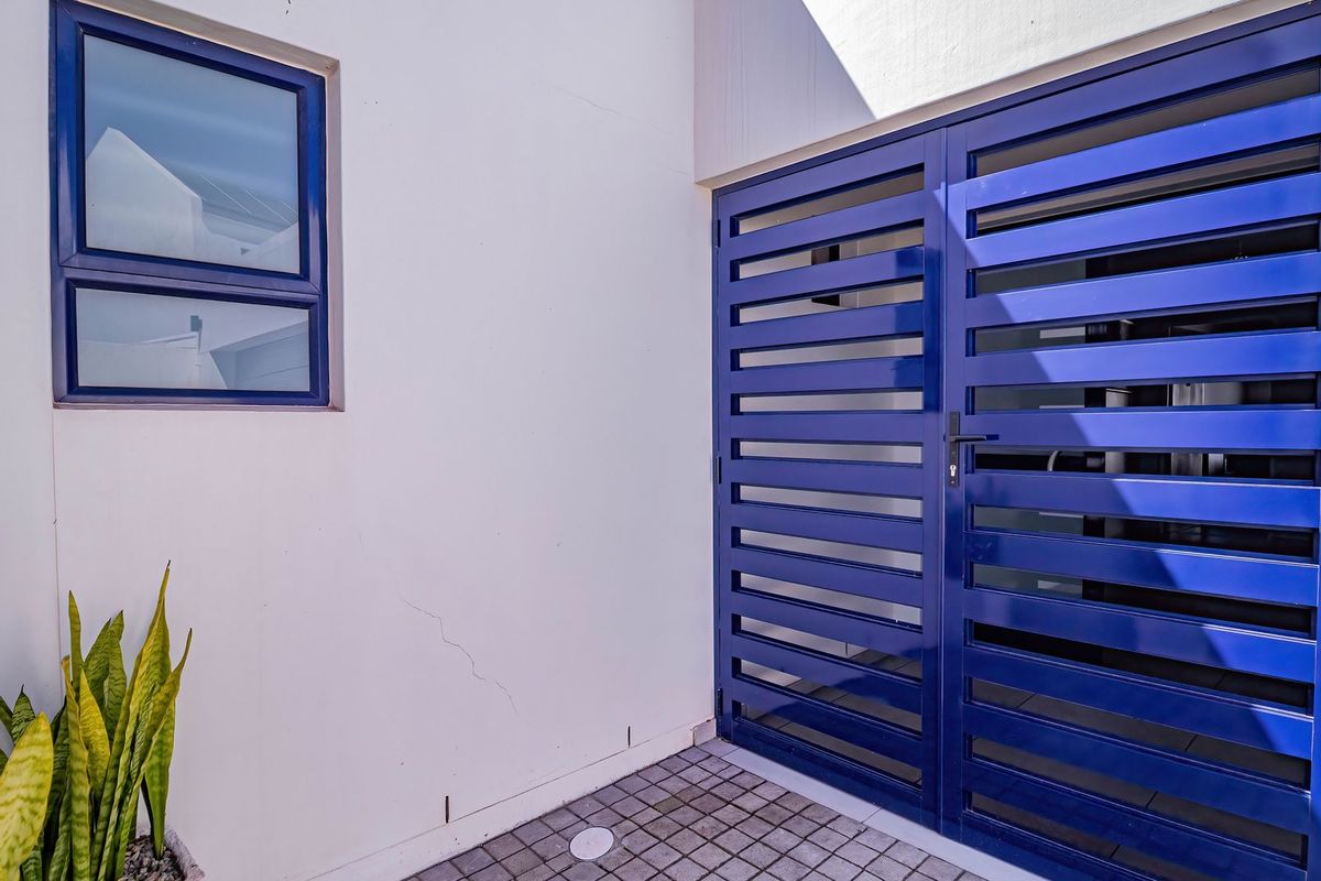 A clean exterior rear area featuring a blue metal security gate, a matching blue-framed window, and paved flooring with a drainage point.