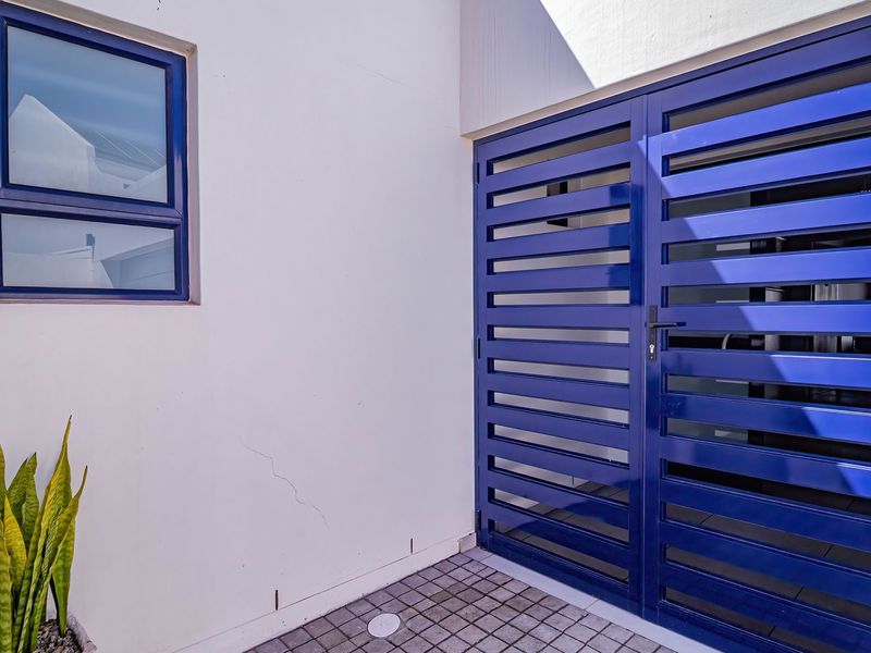 A clean exterior rear area featuring a blue metal security gate, a matching blue-framed window, and paved flooring with a drainage point.