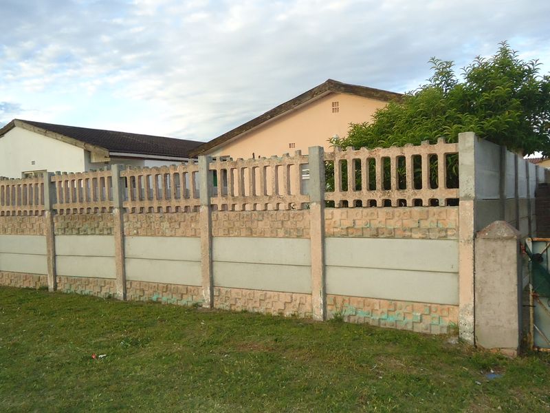 Front view of the property featuring a precast concrete boundary wall and a metal driveway gate, showing signs of weathering and age.