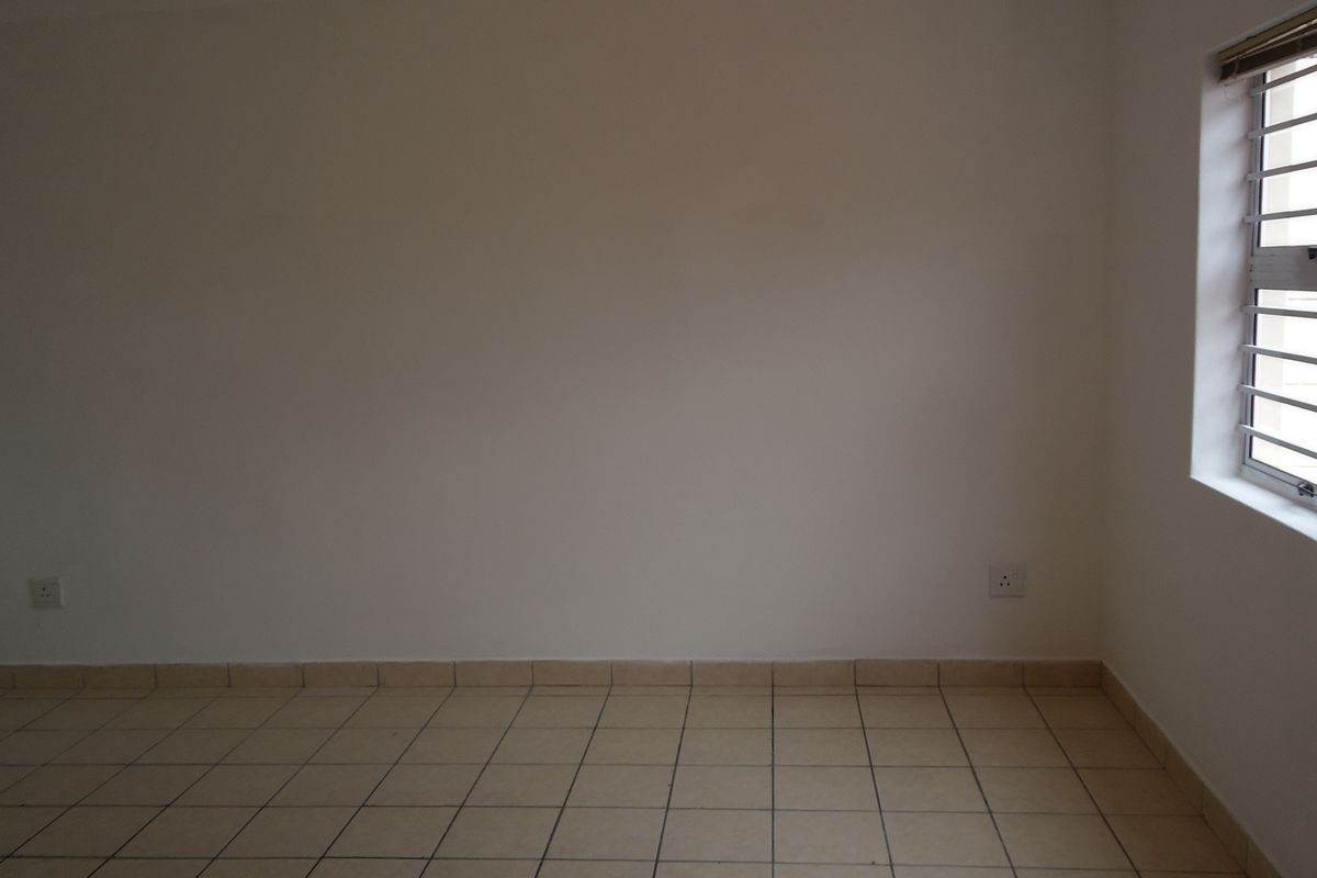 An empty bedroom featuring neutral tiled flooring, plain walls, and a window fitted with security bars.