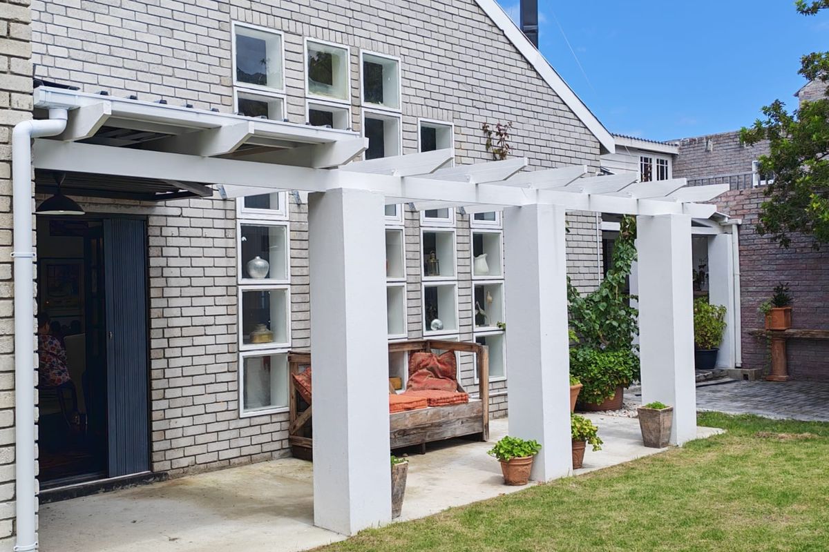 A rear exterior view featuring a white pergola structure, brick facade, and a paved patio area leading to a garden.