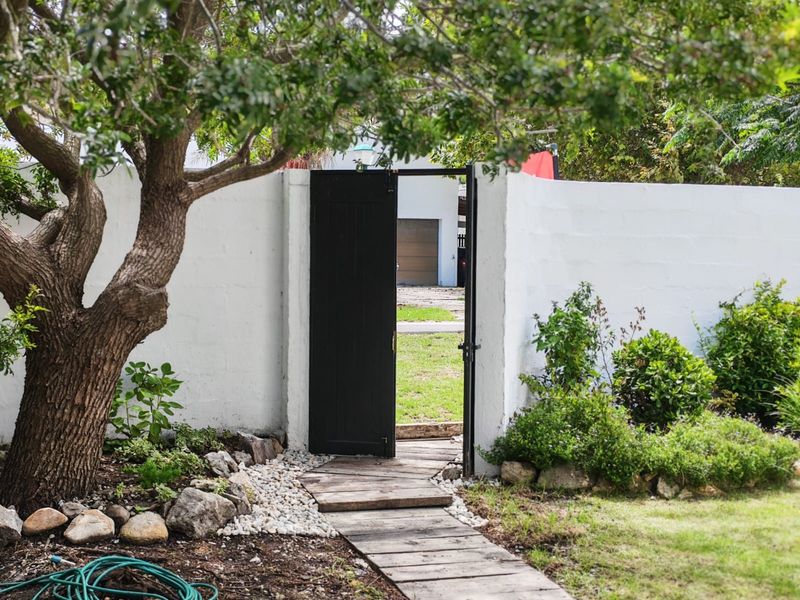 A garden entrance featuring a black gate set within a white boundary wall, complemented by a timber walkway and established landscaping.