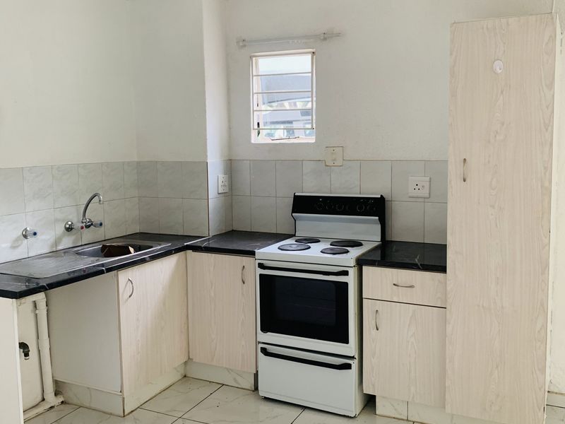 A compact kitchen featuring light wood-finish cabinetry, dark countertops, a freestanding stove, and tiled flooring. The space requires minor repairs to cabinetry and plumbing.