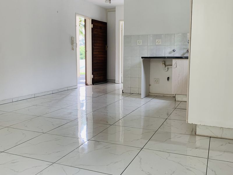 An empty open-plan living and kitchen area featuring white marble-look floor tiles, basic cabinetry, and a tiled splashback.