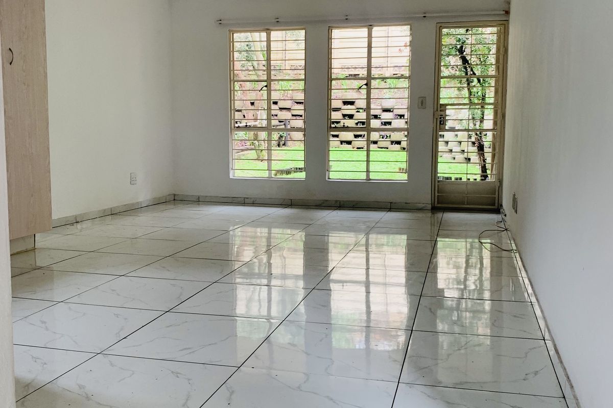 An empty, bright living area featuring polished white marble-look floor tiles, built-in storage, and large windows with security bars overlooking the garden.