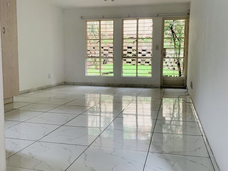 An empty, bright living area featuring polished white marble-look floor tiles, built-in storage, and large windows with security bars overlooking the garden.