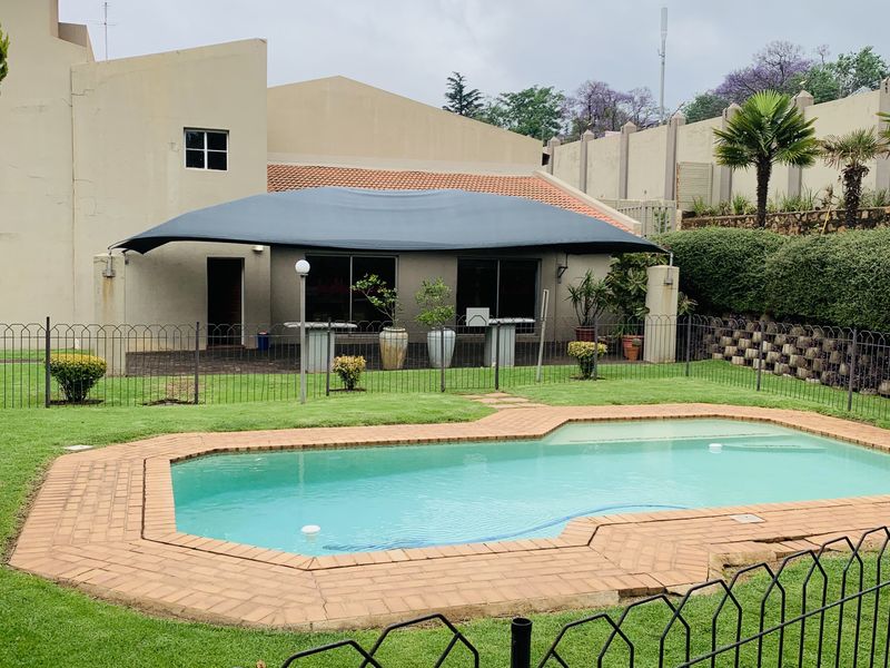 A well-maintained outdoor pool area featuring a swimming pool with brick coping, a protective shade sail, and secure metal perimeter fencing.