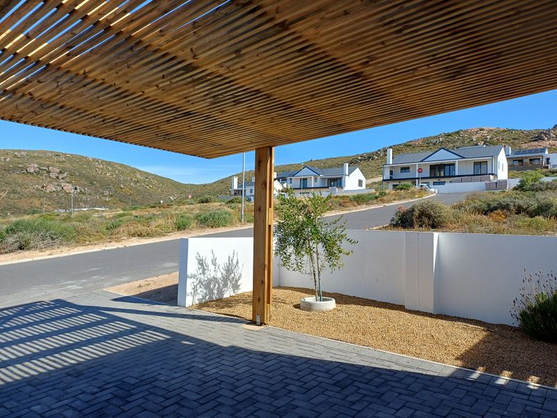 A covered patio area featuring a timber slat pergola, paved flooring, and a low boundary wall, overlooking a landscaped garden and neighbourhood street.