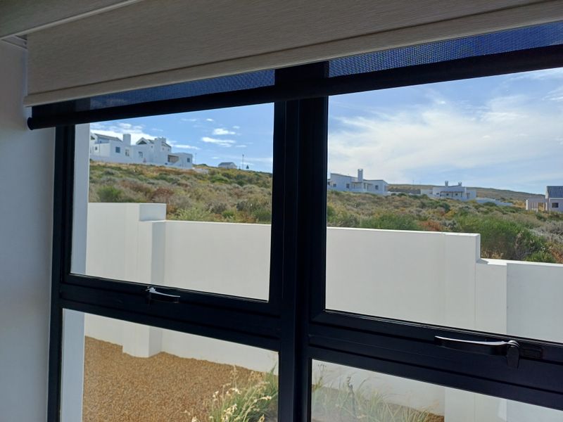 A view through a modern black-framed window looking out onto a landscaped garden area with a white boundary wall and coastal hillside beyond.