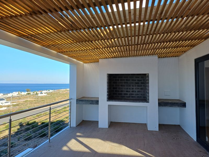 Covered patio featuring a built-in brick braai, wooden slat ceiling, and stainless steel balustrade, offering expansive coastal and landscape views.