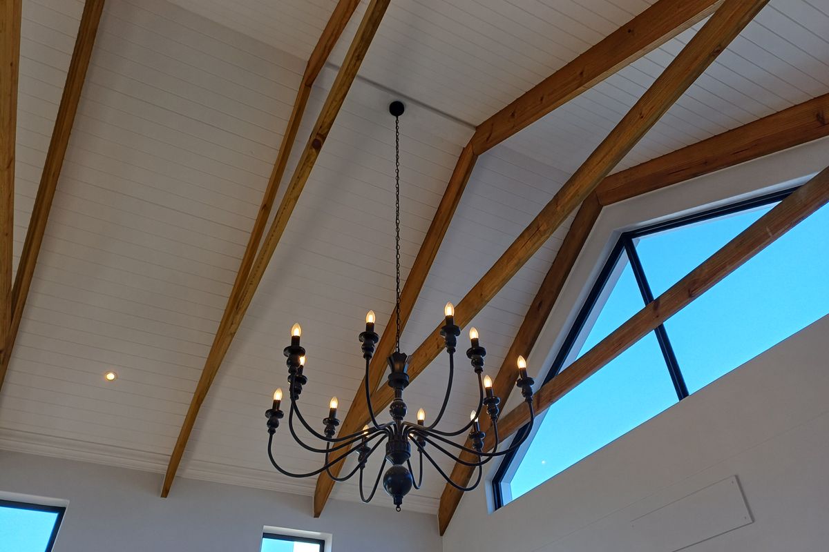 High vaulted ceiling featuring exposed timber beams, tongue-and-groove paneling, and a large feature chandelier, complemented by expansive triangular windows.