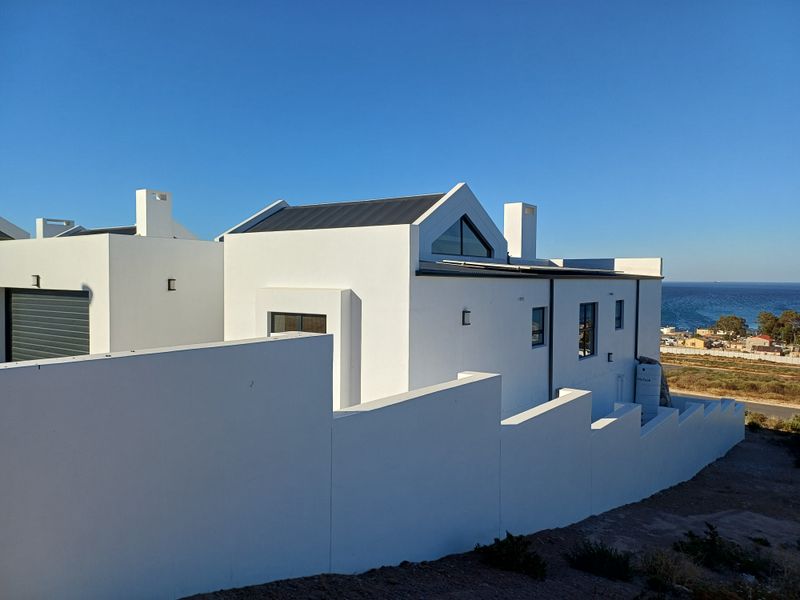 Modern white exterior facade with clean architectural lines, featuring a pitched roof, chimney stacks, and a secure perimeter wall overlooking the coastline.