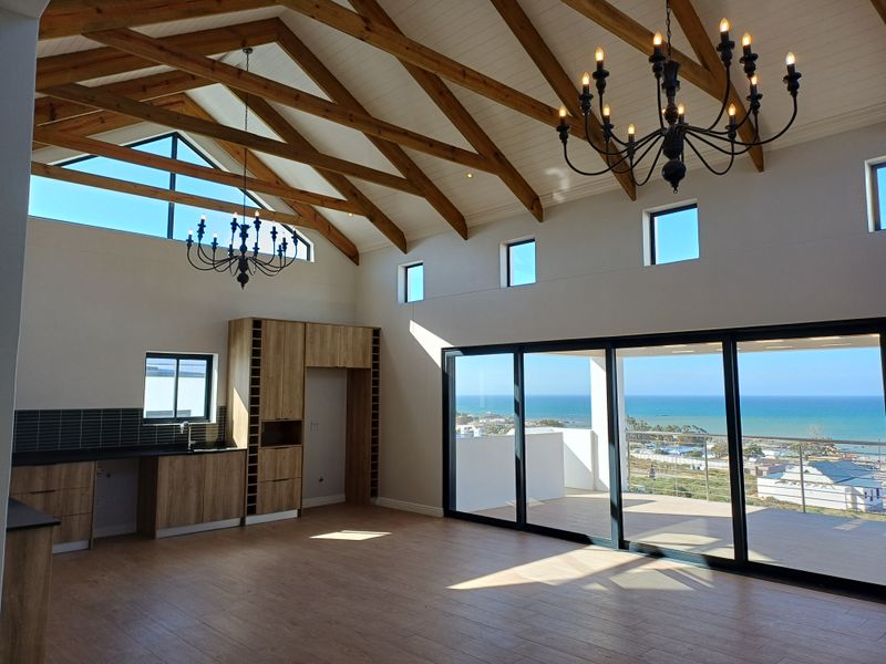 Spacious open-plan living area featuring vaulted timber-beamed ceilings, modern wood-finish cabinetry, and large sliding glass doors opening to a balcony with panoramic ocean views.