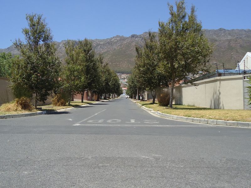 A clear street view of a residential neighborhood featuring paved roads, perimeter boundary walls with electric fencing, and mature trees against a mountain backdrop.