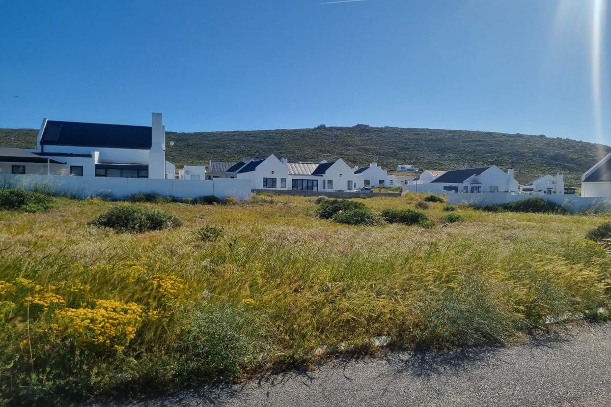 A vacant residential plot featuring natural vegetation, situated in a developed coastal estate with established modern homes in the background.