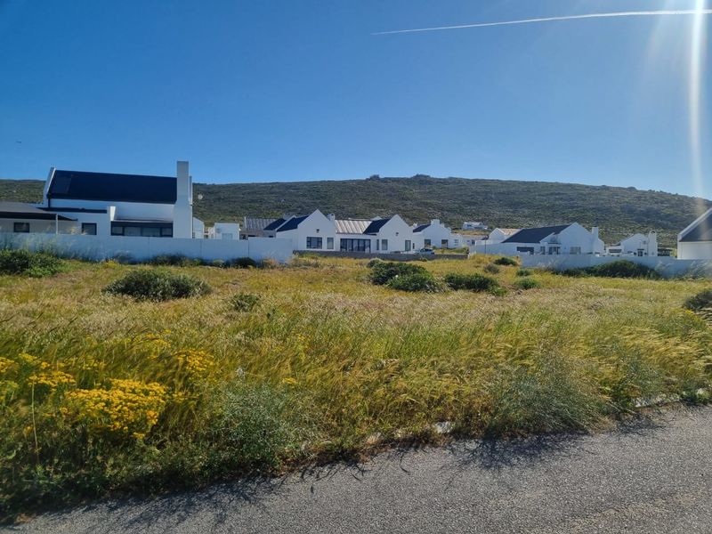 A vacant residential plot featuring natural vegetation, situated in a developed coastal estate with established modern homes in the background.