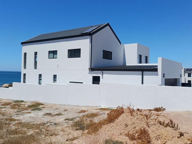 Modern two-story coastal home featuring a clean white facade, dark metal roofing, solar panels, and a secure boundary wall overlooking the ocean.