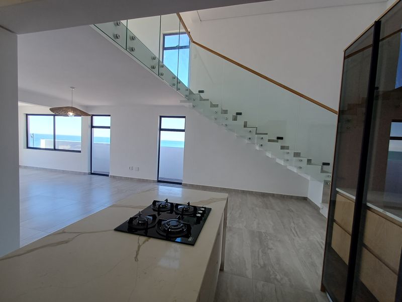 Modern open-plan living area featuring a sleek kitchen island with gas hob and a striking glass balustrade staircase with ocean views.
