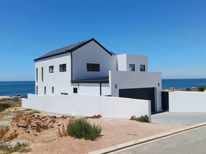 Modern two-story coastal home featuring a clean white facade, dark metal roof, integrated garage, and perimeter boundary walls overlooking the ocean.