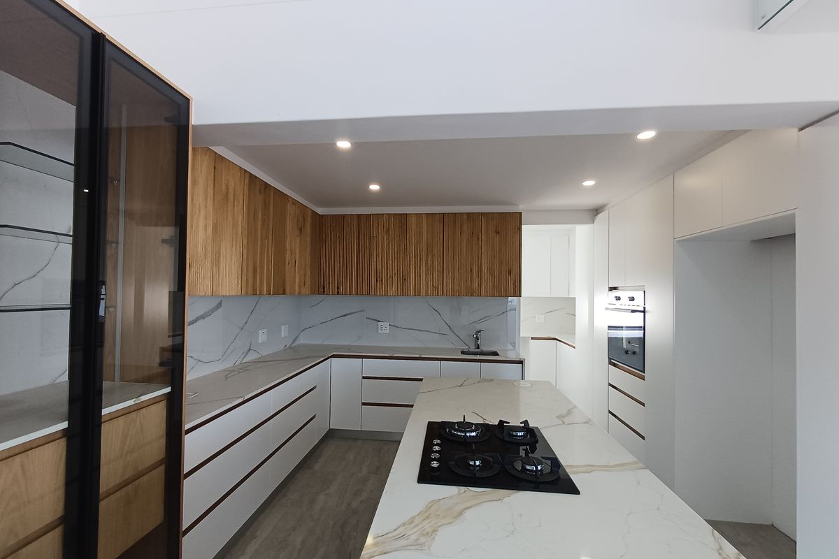 Modern kitchen featuring sleek white and wood-grain cabinetry, marble-look stone countertops, a central island with a gas cooktop, and integrated oven.